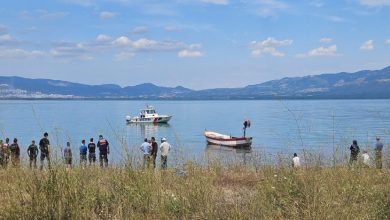 Gölün kıyısında toplanmış insanların karşısında bir sahil güvenlik botu ve küçük bir balıkçı teknesi görülürken, arka planda dağlar ve açık mavi gökyüzü manzarayı tamamlıyor.
