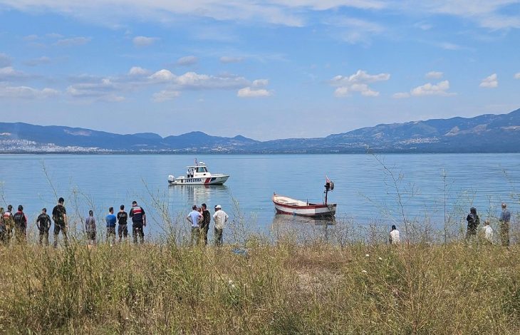 Gölün kıyısında toplanmış insanların karşısında bir sahil güvenlik botu ve küçük bir balıkçı teknesi görülürken, arka planda dağlar ve açık mavi gökyüzü manzarayı tamamlıyor.