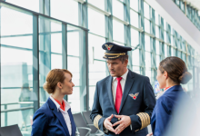 Görselde, havalimanı terminalinde üniformalı bir erkek pilot ile iki kadın uçuş görevlisi ayakta konuşurken görülüyor. Arka planda geniş camlı terminal alanı yer alıyor.