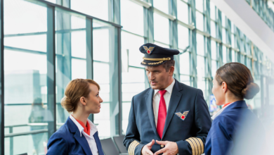 Görselde, havalimanı terminalinde üniformalı bir erkek pilot ile iki kadın uçuş görevlisi ayakta konuşurken görülüyor. Arka planda geniş camlı terminal alanı yer alıyor.