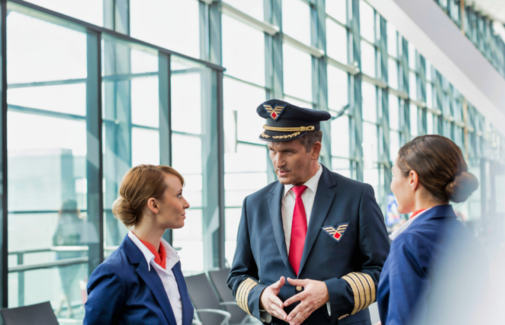Görselde, havalimanı terminalinde üniformalı bir erkek pilot ile iki kadın uçuş görevlisi ayakta konuşurken görülüyor. Arka planda geniş camlı terminal alanı yer alıyor.