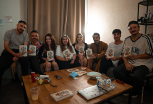 Sıcak ve samimi bir salon ortamında, sekiz kişilik bir ekip bir araya gelmiş şekilde oturuyor. Her biri elinde “Neden Olmasın?” adlı kitabı tutuyor. Grup; koltuk, berjer ve puf gibi farklı oturma alanlarına yerleşmiş, rahat bir sohbet atmosferi içinde görünüyor. Önlerinde ahşap bir sehpa bulunuyor. Sehpada bir dizüstü bilgisayar, bir gazoz kutusu, bir armut, küçük bir mavi kamera ve folyo içinde atıştırmalıklar yer alıyor. Arka planda bej renkli bir perde ve yumuşak ışık veren bir duvar lambası, mekâna sıcak bir ambiyans katıyor. Genel olarak görsel, bir dizi ekibinin kitap etrafında buluştuğu, dayanışma ve paylaşım duygusunu yansıtan samimi bir topluluk fotoğrafı niteliğinde.