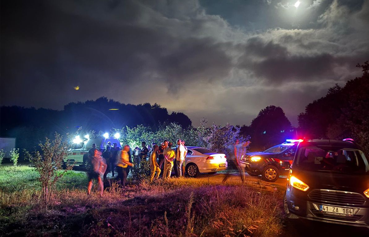 Gece saatlerinde kırsal alanda, ekipler ve vatandaşlar araçların ışıkları altında arama çalışması yürütüyor.