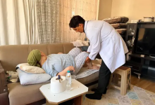 Görselde sağlık görevlisi, ev ortamında koltukta uzanan bir kişiye bacak bölgesinden muayene ve bakım uyguluyor.