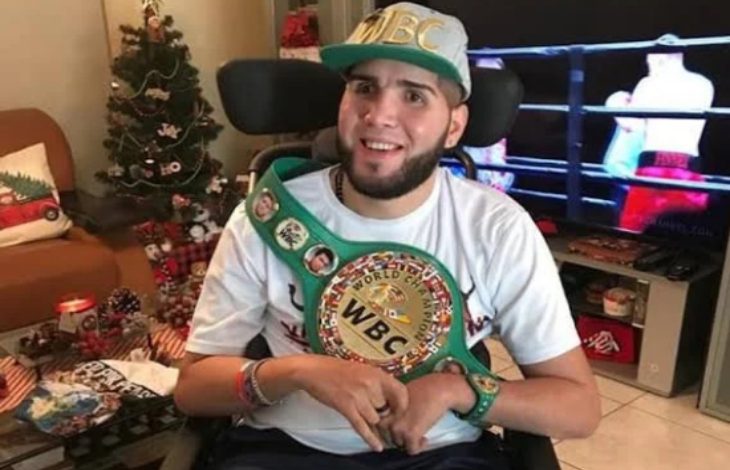 Görselde, boksör Prichard Colón tekerlekli sandalyede otururken kameraya doğru gülümsüyor; üzerinde beyaz bir tişört, başında WBC yazılı bir şapka ve omzunda WBC şampiyonluk kemeri bulunuyor. Arka planda süslenmiş bir Noel ağacı ve televizyonda bir boks karşılaşması görülüyor.