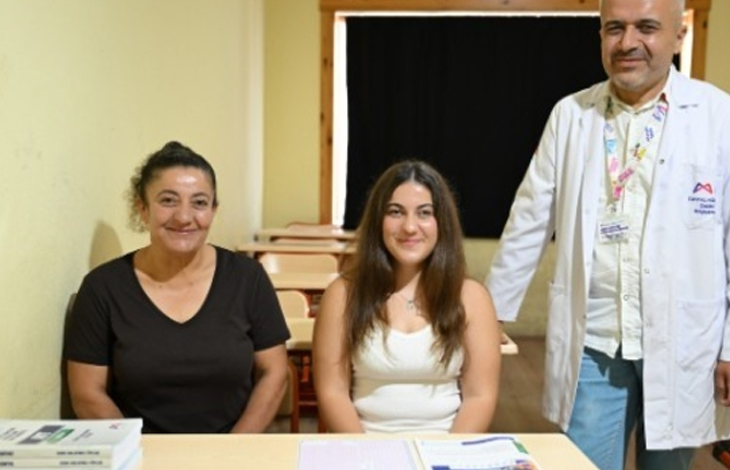 Görselde, bir sınıf ortamında masanın arkasında yan yana oturan iki kadın ile yanlarında beyaz önlükle ayakta duran bir erkek görülüyor. Masanın üzerinde kitaplar ve eğitim materyalleri bulunuyor. Görsel, Mersin’de görme engelli Nisa’nın üniversite hayalinin gerçekleşmesine ilişkin habere ait.