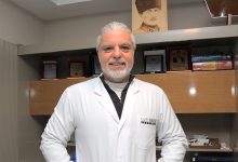 Op. Dr. Alkan Arpaçay, beyaz hekim önlüğüyle, arkasında tıp kitapları, plaketler ve Atatürk portresi bulunan klinik ortamında poz veren bir kalp ve damar cerrahı. Profesyonel, sakin ve güven veren bir duruş sergiliyor.