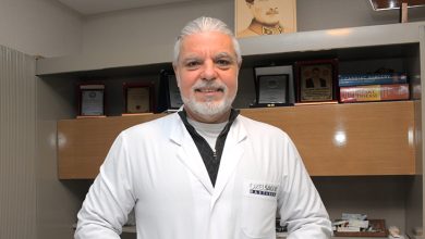 Op. Dr. Alkan Arpaçay, beyaz hekim önlüğüyle, arkasında tıp kitapları, plaketler ve Atatürk portresi bulunan klinik ortamında poz veren bir kalp ve damar cerrahı. Profesyonel, sakin ve güven veren bir duruş sergiliyor.