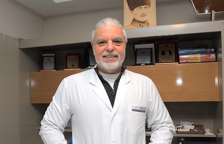Op. Dr. Alkan Arpaçay, beyaz hekim önlüğüyle, arkasında tıp kitapları, plaketler ve Atatürk portresi bulunan klinik ortamında poz veren bir kalp ve damar cerrahı. Profesyonel, sakin ve güven veren bir duruş sergiliyor.