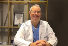 Op. Dr. Ömür Çerçi, beyaz bir hekim önlüğü ve altında mavi cerrahi kıyafetler giymiş şekilde bir masa başında oturuyor. Gözlüklü, gülümseyen ve sakin bir ifadeye sahip. Önlüğünün sol tarafında “EGEP” yazısı bulunuyor. Arka planda raflı bir dolap yer alıyor; raflarda çerçeveli belgeler, kutular ve çeşitli tıbbi ürünler görülüyor. Ortam bir hastane ya da klinik ofisi izlenimi veriyor.