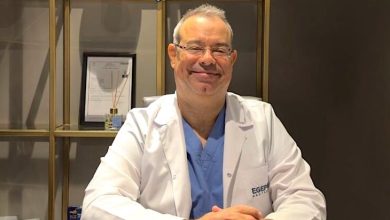 Op. Dr. Ömür Çerçi, beyaz bir hekim önlüğü ve altında mavi cerrahi kıyafetler giymiş şekilde bir masa başında oturuyor. Gözlüklü, gülümseyen ve sakin bir ifadeye sahip. Önlüğünün sol tarafında “EGEP” yazısı bulunuyor. Arka planda raflı bir dolap yer alıyor; raflarda çerçeveli belgeler, kutular ve çeşitli tıbbi ürünler görülüyor. Ortam bir hastane ya da klinik ofisi izlenimi veriyor.