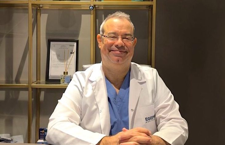 Op. Dr. Ömür Çerçi, beyaz bir hekim önlüğü ve altında mavi cerrahi kıyafetler giymiş şekilde bir masa başında oturuyor. Gözlüklü, gülümseyen ve sakin bir ifadeye sahip. Önlüğünün sol tarafında “EGEP” yazısı bulunuyor. Arka planda raflı bir dolap yer alıyor; raflarda çerçeveli belgeler, kutular ve çeşitli tıbbi ürünler görülüyor. Ortam bir hastane ya da klinik ofisi izlenimi veriyor.