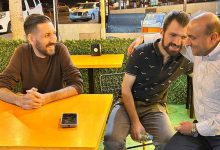 Üç kişinin sarı masalarda oturduğu, akşam saatlerini andıran dış mekân kafesinde samimi bir sohbet anı görülüyor. İki kişi yan yana oturup yakın bir poz verirken, diğer kişi karşı masada telefonunun yanında oturuyor. Arkada yapay yeşillik, park hâlindeki araçlar ve sokak ışıkları bulunuyor.