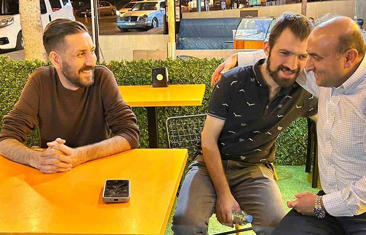 Üç kişinin sarı masalarda oturduğu, akşam saatlerini andıran dış mekân kafesinde samimi bir sohbet anı görülüyor. İki kişi yan yana oturup yakın bir poz verirken, diğer kişi karşı masada telefonunun yanında oturuyor. Arkada yapay yeşillik, park hâlindeki araçlar ve sokak ışıkları bulunuyor.