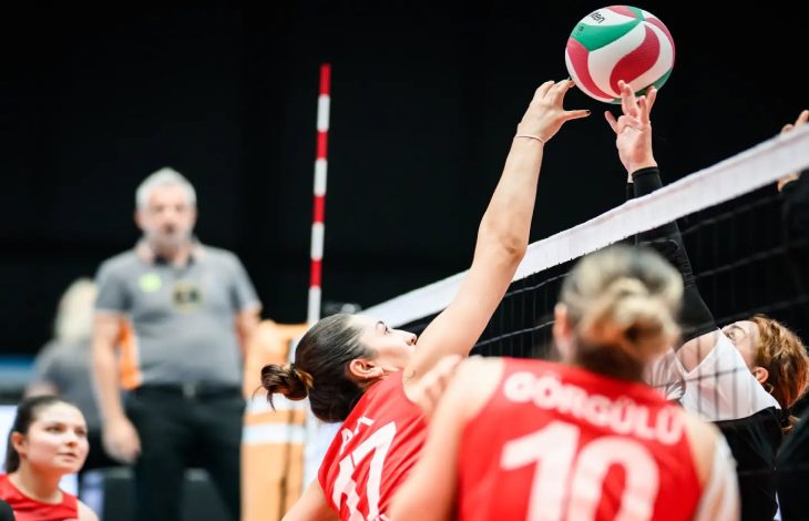 Görselde oturarak voleybol kadınlar müsabakasından heyecanlı bir an görülüyor. Kırmızı forma giyen Türkiye takımı oyuncuları, file önünde rakipleriyle mücadele ediyor. Ön planda sırtında “Türkiye” yazısı ve 10 numarası bulunan bir millî takım oyuncusu dikkat çekiyor. Ortada ise kırmızı formalı başka bir oyuncu, topa müdahale etmek için fileye doğru uzanmış durumda. Rakip takımın oyuncuları da file üzerinde topa karşı hamle yapıyor.