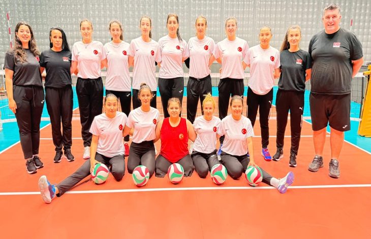 Görselde, kapalı bir spor salonunda Türk kadın oturarak voleybol milli takımının oyuncuları ve teknik ekibi, voleybol toplarıyla birlikte takım fotoğrafı için poz verirken görülüyor.