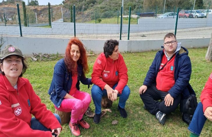 Özel gereksinimli gençler ve yanlarında bulunan ebeveyn kadın yeşillik bir alanda oturuyor.