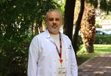Prof. Dr. Ünal Aydın, açık havada, ağaçlar ve yeşilliklerle çevrili bir yürüyüş yolunda duruyor. Üzerinde beyaz hekim önlüğü, altında bej tonlarında bir gömlek bulunuyor. Boynunda bordo renkli bir yaka kartı askısı ve kırmızı bir klips dikkat çekiyor. Arka planda hem gölgeli hem güneş alan bölgeler, uzakta park hâlinde araçlar görülüyor. Aydın’ın duruşu ve ortam, kendisini profesyonel, güven veren ve sahada aktif çalışan bir cerrah olarak yansıtıyor.