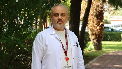 Prof. Dr. Ünal Aydın, açık havada, ağaçlar ve yeşilliklerle çevrili bir yürüyüş yolunda duruyor. Üzerinde beyaz hekim önlüğü, altında bej tonlarında bir gömlek bulunuyor. Boynunda bordo renkli bir yaka kartı askısı ve kırmızı bir klips dikkat çekiyor. Arka planda hem gölgeli hem güneş alan bölgeler, uzakta park hâlinde araçlar görülüyor. Aydın’ın duruşu ve ortam, kendisini profesyonel, güven veren ve sahada aktif çalışan bir cerrah olarak yansıtıyor.