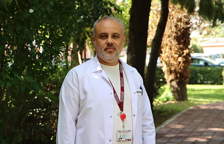 Prof. Dr. Ünal Aydın, açık havada, ağaçlar ve yeşilliklerle çevrili bir yürüyüş yolunda duruyor. Üzerinde beyaz hekim önlüğü, altında bej tonlarında bir gömlek bulunuyor. Boynunda bordo renkli bir yaka kartı askısı ve kırmızı bir klips dikkat çekiyor. Arka planda hem gölgeli hem güneş alan bölgeler, uzakta park hâlinde araçlar görülüyor. Aydın’ın duruşu ve ortam, kendisini profesyonel, güven veren ve sahada aktif çalışan bir cerrah olarak yansıtıyor.