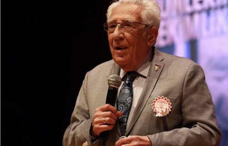 Prof. Dr. Coşkun Özdemir bir sahnede, elinde mikrofonla konuşma yaparken görülüyor. Üzerinde açık gri bir ceket, beyaz gömlek ve desenli mavi bir kravat bulunuyor. Ceketinin sol tarafında kırmızı kenarlı yuvarlak bir rozet takılı. Arka planda, bulanık şekilde görünen bir afiş ya da projeksiyon alanı var; bu da ortamın bir tören, konferans veya anma programı olduğunu düşündürüyor. Özdemir’in duruşu sakin, kendinden emin ve saygın bir konuşmacı izlenimi veriyor.