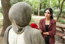 Görselde açık alanda yapılan bir röportaj sırasında, başörtülü bir kadın sırtı kameraya dönük şekilde dururken karşısındaki kadın muhabir mikrofon uzatıyor.