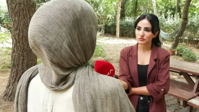 Görselde açık alanda yapılan bir röportaj sırasında, başörtülü bir kadın sırtı kameraya dönük şekilde dururken karşısındaki kadın muhabir mikrofon uzatıyor.