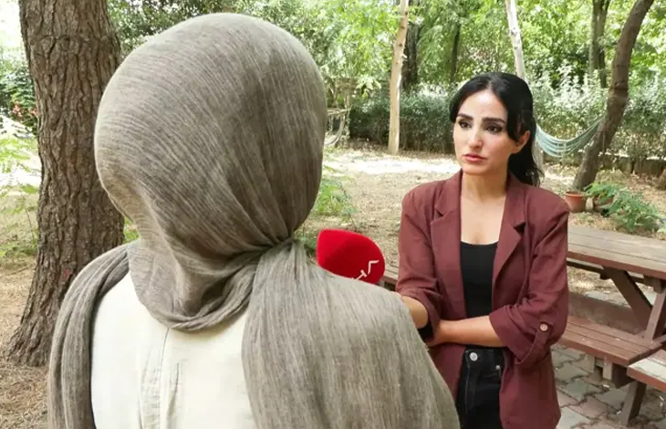 Görselde açık alanda yapılan bir röportaj sırasında, başörtülü bir kadın sırtı kameraya dönük şekilde dururken karşısındaki kadın muhabir mikrofon uzatıyor.