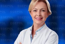 Kısa açık renk saçlı ve gülümseyen Uzm. Dr. Tuğba Emüroğlu, beyaz hekim önlüğüyle kollarını çapraz şekilde bağlamış halde görülüyor. Arka planda mavi zemin üzerinde tekrarlanan “EGEPOL HASTANELERİ” yazıları yer alıyor. Genel görünüm profesyonel, güven veren ve kurumsal bir sağlık ortamını yansıtıyor.