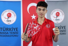 Görselde, Ebubekir İnce kırmızı Türkiye formasıyla elinde masa tenisi raketi ve top tutarak kameraya bakıyor. Arka planda Türk bayrağı ile Türkiye Masa Tenisi Federasyonu logolarının bulunduğu panolar yer alıyor.