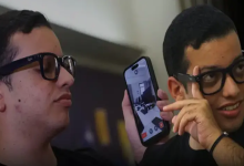 Görselde, yan yana duran iki gençten biri elinde tuttuğu akıllı telefonun ekranını gösteriyor. Her iki genç de koyu renkli gözlük takıyor.