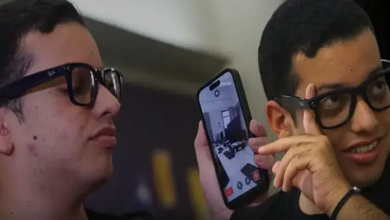 Görselde, yan yana duran iki gençten biri elinde tuttuğu akıllı telefonun ekranını gösteriyor. Her iki genç de koyu renkli gözlük takıyor.