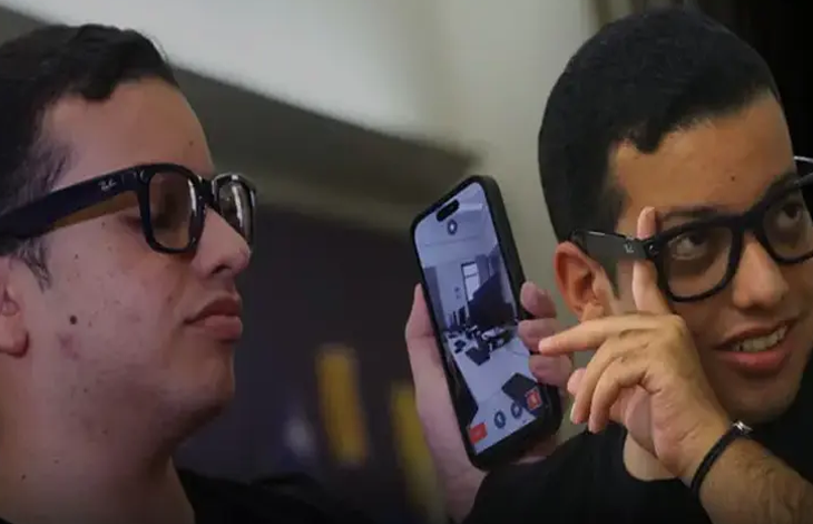 Görselde, yan yana duran iki gençten biri elinde tuttuğu akıllı telefonun ekranını gösteriyor. Her iki genç de koyu renkli gözlük takıyor.