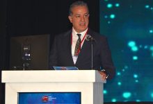 İZDO Yönetim Kurulu Başkanı Ersin Atinel bir kürsü arkasında konuşma yaparken görülüyor. Üzerinde koyu renk takım elbise, beyaz gömlek ve desenli bir kravat bulunuyor. Boynunda kırmızı bir yaka kartı şeridi yer alıyor. Konuştuğu kürsünün ön yüzünde İzmir Dişhekimleri Odası logosu ve yazısı bulunan bir ekran bulunuyor. Arka planda, sahneyi aydınlatan mavi-yeşil tonlarda dijital ışık desenleri yer alıyor. Genel atmosfer, resmi bir toplantı, panel veya bilimsel etkinlik izlenimi veriyor.