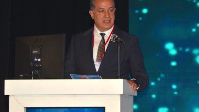 İZDO Yönetim Kurulu Başkanı Ersin Atinel bir kürsü arkasında konuşma yaparken görülüyor. Üzerinde koyu renk takım elbise, beyaz gömlek ve desenli bir kravat bulunuyor. Boynunda kırmızı bir yaka kartı şeridi yer alıyor. Konuştuğu kürsünün ön yüzünde İzmir Dişhekimleri Odası logosu ve yazısı bulunan bir ekran bulunuyor. Arka planda, sahneyi aydınlatan mavi-yeşil tonlarda dijital ışık desenleri yer alıyor. Genel atmosfer, resmi bir toplantı, panel veya bilimsel etkinlik izlenimi veriyor.