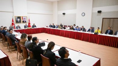 Geniş ve modern bir toplantı salonunda, U şeklinde düzenlenmiş masaların etrafında çok sayıda kişinin toplantı yaptığı, masaların üzerinde mikrofon, su şişeleri ve belgelerin bulunduğu görülmektedir.