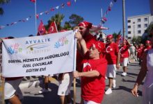 Ayvalık Belediyesi Sosyal Hizmetler Müdürlüğü Özel Çocuklar Eğitim Evi yazılı büyük bir pankartı taşıyarak sokakta coşkuyla yürüyen kırmızı tişörtlü ve beyaz şortlu bando veya geçit töreni grubu yer alıyor.