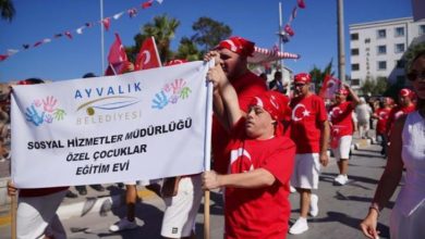 Ayvalık Belediyesi Sosyal Hizmetler Müdürlüğü Özel Çocuklar Eğitim Evi yazılı büyük bir pankartı taşıyarak sokakta coşkuyla yürüyen kırmızı tişörtlü ve beyaz şortlu bando veya geçit töreni grubu yer alıyor.