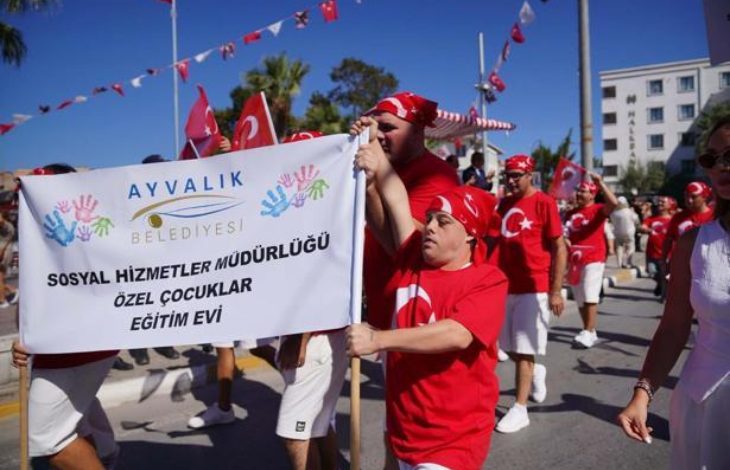Ayvalık Belediyesi Sosyal Hizmetler Müdürlüğü Özel Çocuklar Eğitim Evi yazılı büyük bir pankartı taşıyarak sokakta coşkuyla yürüyen kırmızı tişörtlü ve beyaz şortlu bando veya geçit töreni grubu yer alıyor.