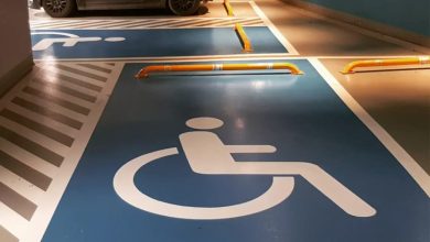 Kapalı bir otopark alanı görülüyor. Zeminde mavi renkle belirlenmiş engelli park yerleri bulunuyor ve her birinin üzerinde beyaz tekerlekli sandalye sembolleri yer alıyor. Park yerlerinin önünde araçların ileri gitmesini engelleyen sarı bariyerler var. Arka tarafta koyu renkli bir araç bu alanlardan birine park etmiş durumda. Mekân temiz, düzenli ve erişilebilirlik işaretlemeleri belirgin.
