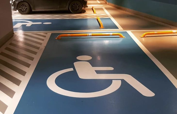 Kapalı bir otopark alanı görülüyor. Zeminde mavi renkle belirlenmiş engelli park yerleri bulunuyor ve her birinin üzerinde beyaz tekerlekli sandalye sembolleri yer alıyor. Park yerlerinin önünde araçların ileri gitmesini engelleyen sarı bariyerler var. Arka tarafta koyu renkli bir araç bu alanlardan birine park etmiş durumda. Mekân temiz, düzenli ve erişilebilirlik işaretlemeleri belirgin.