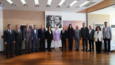 Modern bir toplantı salonunda yan yana dizilmiş kalabalık bir protokol grubu görülüyor. Ortada Aile Bakanı, yanında Aydın Valisi ve Aydın Belediye Başkanı olmak üzere toplamda on üç kişi resmi bir poz veriyor. Her biri takım elbise ve profesyonel kıyafetlerle, arka planda ise duvarda asılı iki adet Atatürk portresi bulunuyor. Ortam resmi, düzenli ve protokol buluşması niteliğinde.