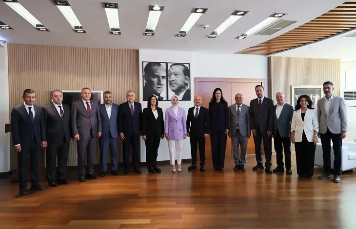 Modern bir toplantı salonunda yan yana dizilmiş kalabalık bir protokol grubu görülüyor. Ortada Aile Bakanı, yanında Aydın Valisi ve Aydın Belediye Başkanı olmak üzere toplamda on üç kişi resmi bir poz veriyor. Her biri takım elbise ve profesyonel kıyafetlerle, arka planda ise duvarda asılı iki adet Atatürk portresi bulunuyor. Ortam resmi, düzenli ve protokol buluşması niteliğinde.