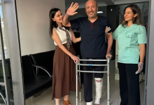 Hastane veya klinik koridorunda, yürüteç (walker) desteğiyle ayakta duran ve sol bacağı dize kadar beyaz sargılar içinde olan orta yaşlı bir erkek hasta görülüyor. Koyu mavi kıyafetli hasta gülümseyerek sağ elini havaya kaldırıp el sallarken, sol tarafında kahverengi etekli ve beyaz bluzlu bir yakını, sağ tarafında ise açık yeşil medikal forma giymiş kadın sağlık çalışanı ona destek vererek mutlulukla gülümsüyor.