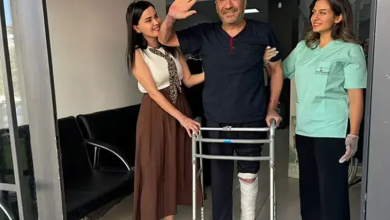 Hastane veya klinik koridorunda, yürüteç (walker) desteğiyle ayakta duran ve sol bacağı dize kadar beyaz sargılar içinde olan orta yaşlı bir erkek hasta görülüyor. Koyu mavi kıyafetli hasta gülümseyerek sağ elini havaya kaldırıp el sallarken, sol tarafında kahverengi etekli ve beyaz bluzlu bir yakını, sağ tarafında ise açık yeşil medikal forma giymiş kadın sağlık çalışanı ona destek vererek mutlulukla gülümsüyor.