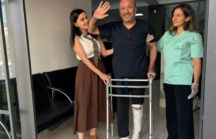 Hastane veya klinik koridorunda, yürüteç (walker) desteğiyle ayakta duran ve sol bacağı dize kadar beyaz sargılar içinde olan orta yaşlı bir erkek hasta görülüyor. Koyu mavi kıyafetli hasta gülümseyerek sağ elini havaya kaldırıp el sallarken, sol tarafında kahverengi etekli ve beyaz bluzlu bir yakını, sağ tarafında ise açık yeşil medikal forma giymiş kadın sağlık çalışanı ona destek vererek mutlulukla gülümsüyor.