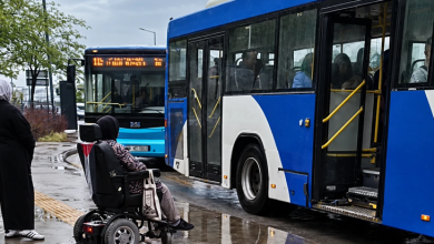 Görselin üst bölümünde, yağmurlu ve ıslak bir havada durakta bekleyen akülü tekerlekli sandalyeli bir birey ile mavi-beyaz renkli belediye otobüsleri yer alıyor. Sağ tarafta kapıları açık olan otobüsün basamak hizasında duran açılmamış engelli rampası görülürken, ön planda arkası dönük oturan engelli birey araca binemeden durakta bekliyor. Sol tarafta siyah giyimli bir yaya, sarı hissedilebilir görme engelli kılavuz çizgileri, yerdeki yağmur birikintileri ve geride bekleyen bir diğer EGO otobüsü ile bulutlu gökyüzü seçiliyor.