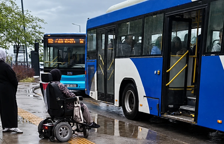 Görselin üst bölümünde, yağmurlu ve ıslak bir havada durakta bekleyen akülü tekerlekli sandalyeli bir birey ile mavi-beyaz renkli belediye otobüsleri yer alıyor. Sağ tarafta kapıları açık olan otobüsün basamak hizasında duran açılmamış engelli rampası görülürken, ön planda arkası dönük oturan engelli birey araca binemeden durakta bekliyor. Sol tarafta siyah giyimli bir yaya, sarı hissedilebilir görme engelli kılavuz çizgileri, yerdeki yağmur birikintileri ve geride bekleyen bir diğer EGO otobüsü ile bulutlu gökyüzü seçiliyor.