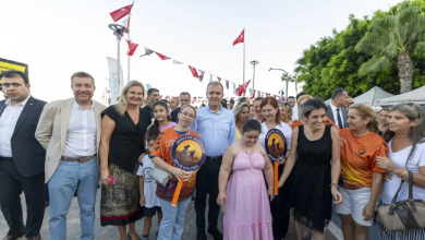 Görselde Mersin Büyükşehir Belediye Başkanı, engelli gençler ve aileleriyle birlikte açık alanda toplu fotoğraf çekiminde görülüyor. Başkan, grubun ortasında yer alıyor; etrafında turuncu tişörtleriyle “Engelliler Spor Kulübü” üyeleri ve aileleri bulunuyor. Katılımcıların bir kısmı kulübün logosunun yer aldığı yuvarlak pankartları taşıyor. Arka planda Türk bayrakları, ağaçlar ve etkinlik çadırları yer alıyor. Atmosfer sıcak, destekleyici ve toplumsal katılım vurgusuyla bir etkinlik ortamını yansıtıyor.