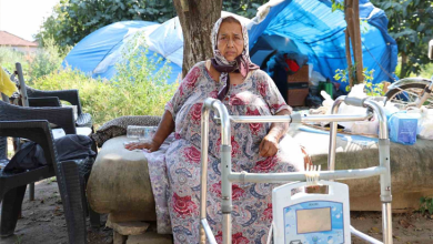 Görselin üst bölümünde, açık alanda bir ağacın gölgesindeki sedire oturmuş olan Leyla Demirkafa yer alıyor. Başörtülü ve desenli geniş bir elbise giymiş olan kadın, ciddi ve yorgun bir yüz ifadesiyle objektife bakıyor. Hemen önünde tutunarak ayağa kalkmasını sağlayan metal bir yürüteç (walker) ve üzerinde asılı duran tıbbi bir cihaz bulunuyor. Çevresinde plastik sandalyeler, arka planda ise mavi naylon brandalarla oluşturulmuş çadır benzeri barınma alanları ve yeşil ağaçlar görülüyor.